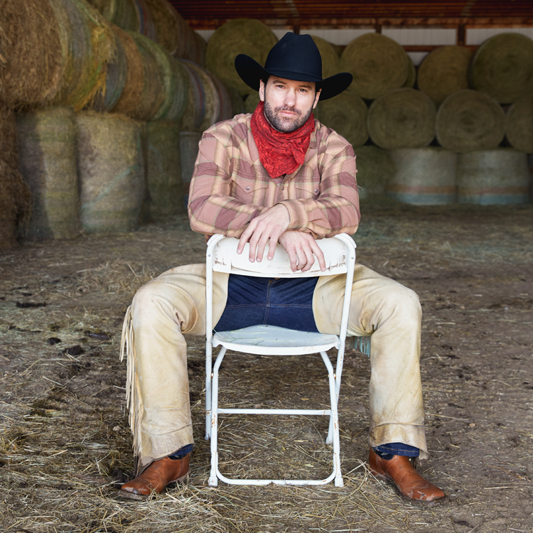 A man wearing chaps dressed like a cowboy sitting in a chair in the hay barn.